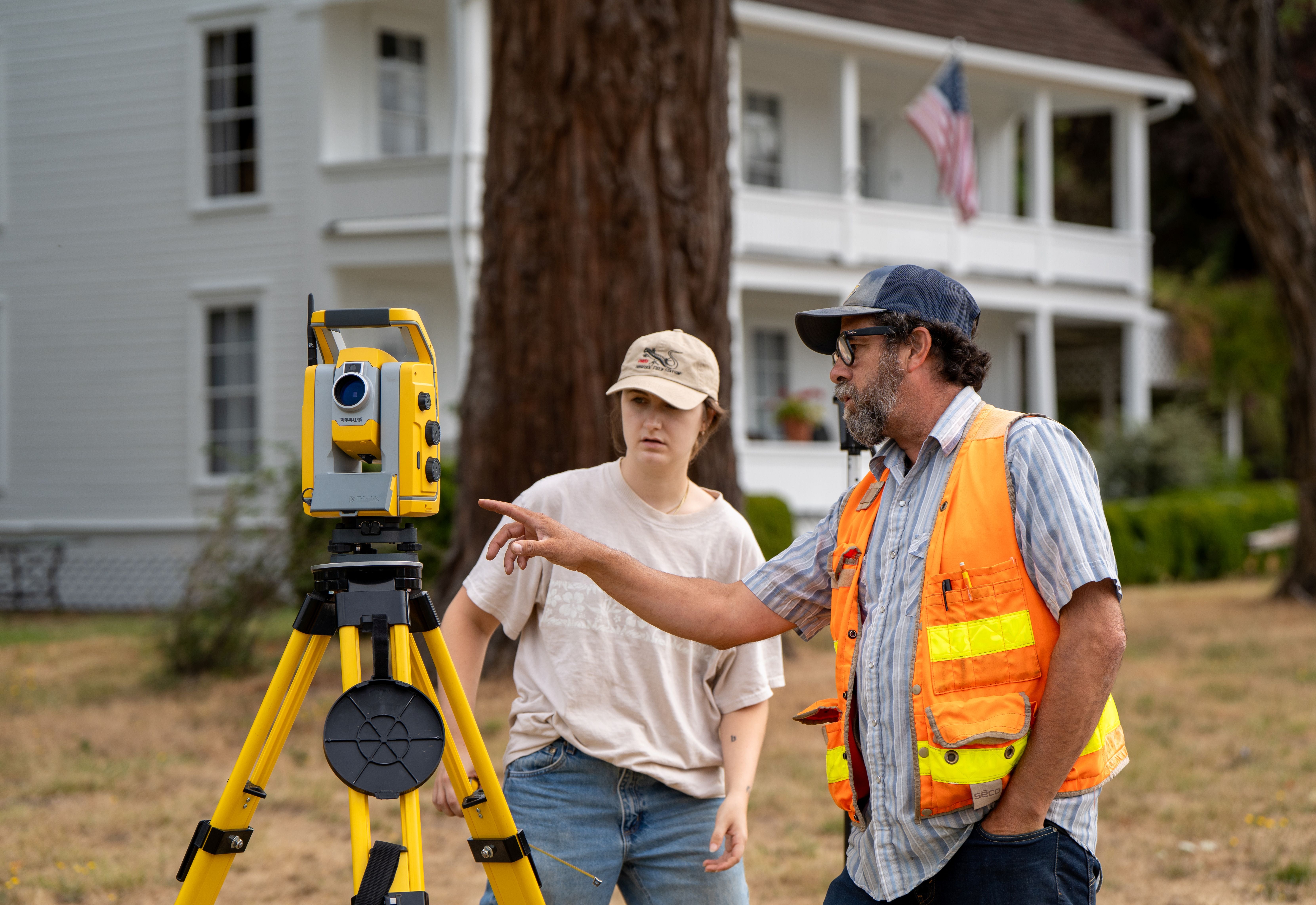 An instructor wearing a high visibility vest shows a student something on a total station. In the background of the photo is the historic Applegate family house. 