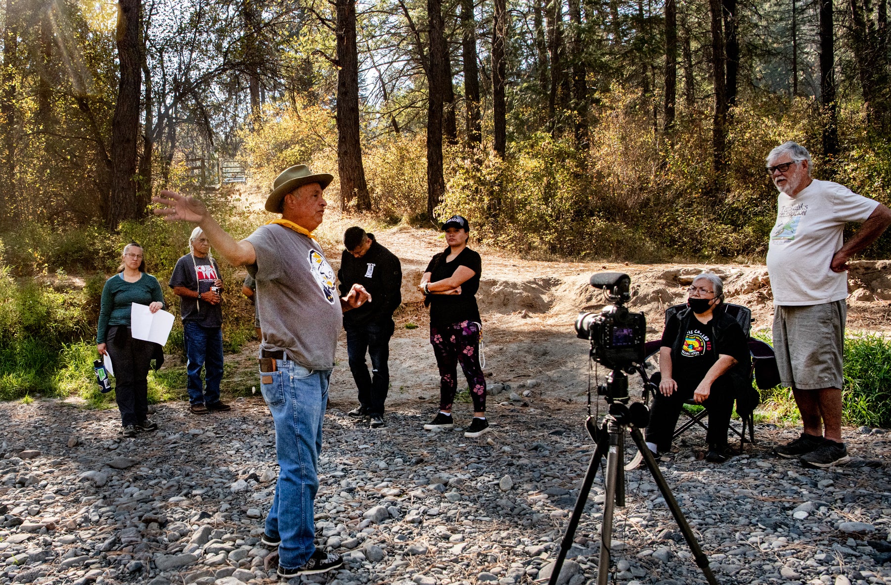 An Indigenous man tells a story to a group of Indigenous and white people in a forest. A camera is filming him.