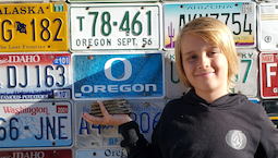 a young boy with long blond hair holds a mammoth tooth against a wall of license plates