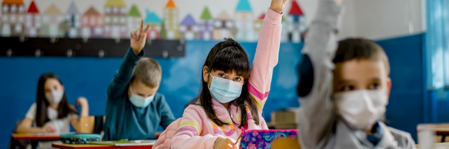 Students wearing masks in a classroom, many of whom are raising their hands