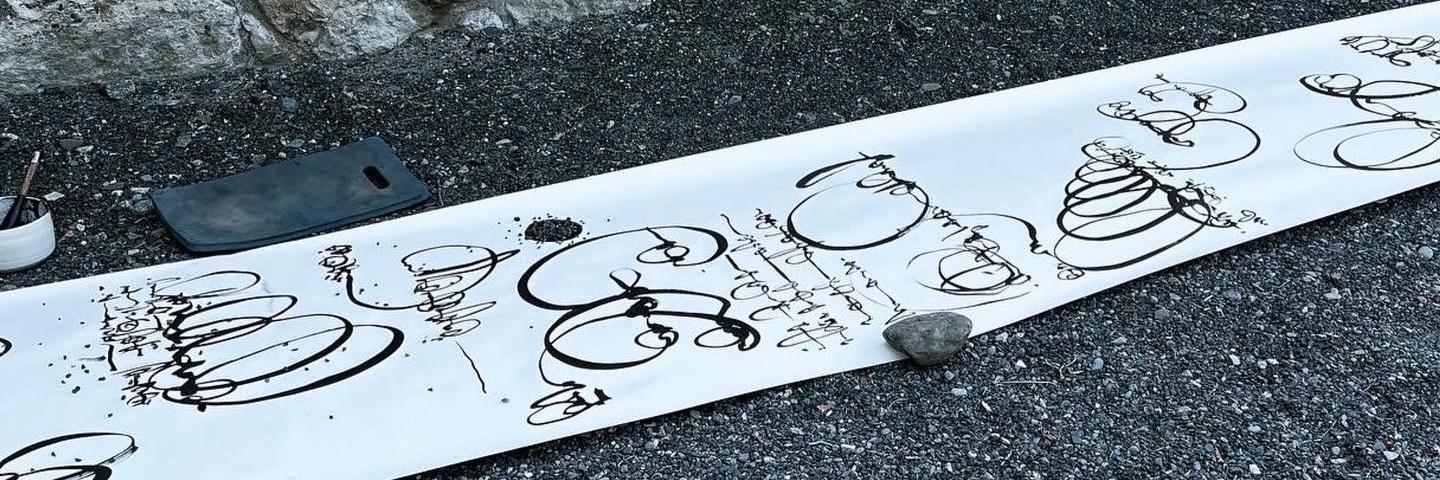 A long scroll of paper on the dark gravel ground. It's held down by normal rocks and is covered in calligraphic swirls of dark ink. 