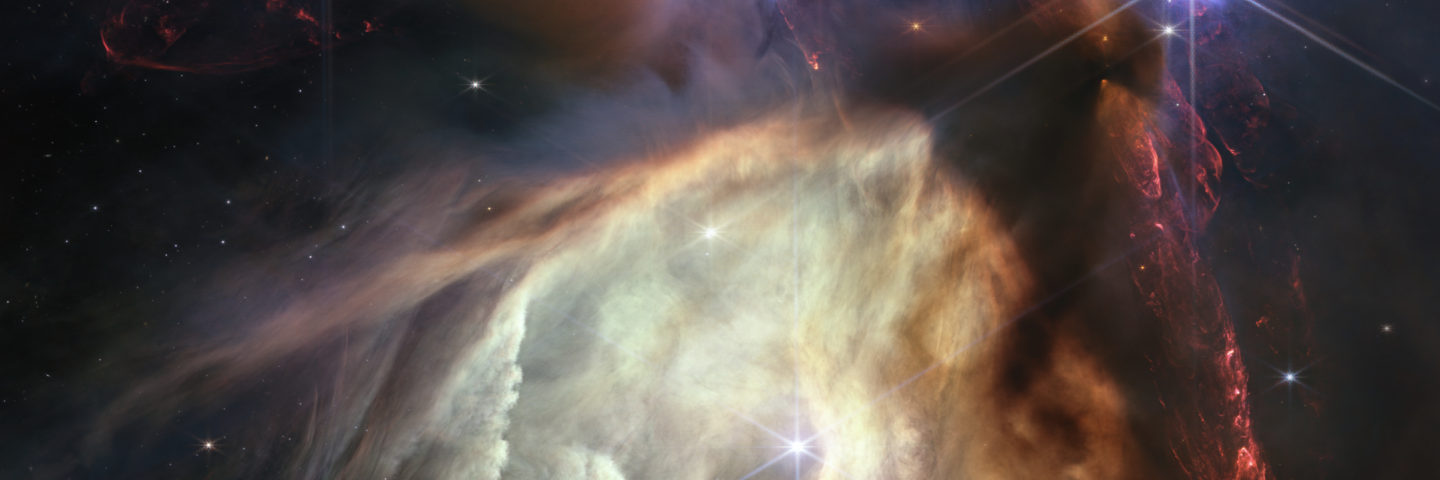 Red dual opposing jets coming from young stars fill the darker top half of the image, while a glowing pale-yellow, cave-like structure is bottom center, tilted toward two o’clock, with a bright star at its center. 