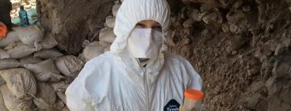 Person in full suite with a trowel and collection cups in hands.