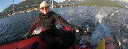 Sarah Gravem, wearing sunglasses and a white hat, pilots a motorboat in a body of water with a bridge and mountains behind her.