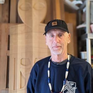 Greg Robinson stands in front of some of his wood carving and looks at the camera