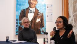 A Black woman speaks into a microphone as a white woman looks on.