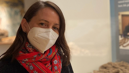 White woman wearing a red scarf and a white mask stands in front of a museum case featuring a net made of natural fibers